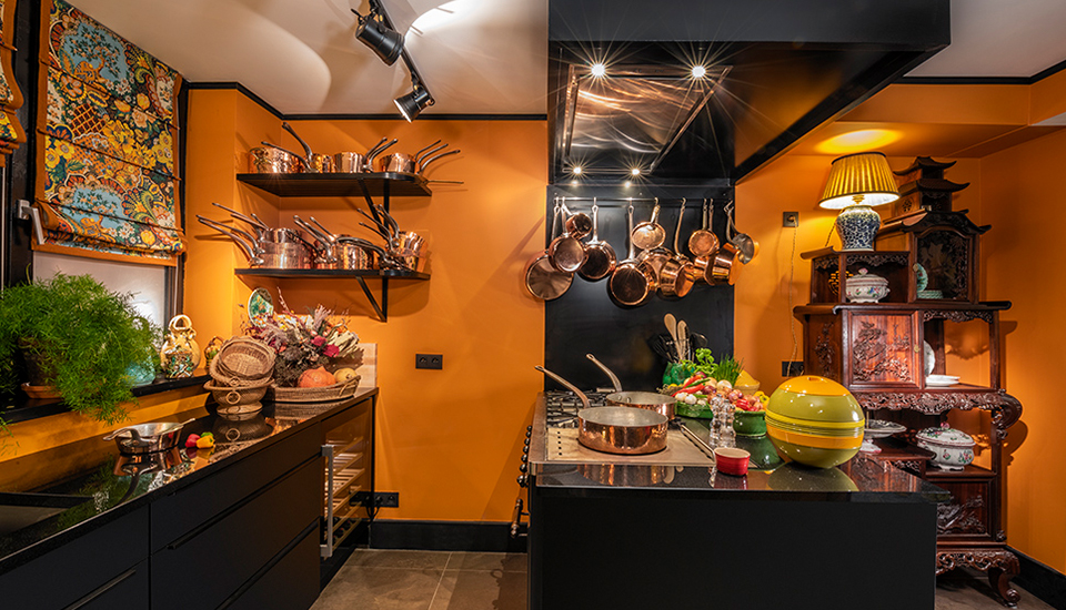 Cuisine avec murs orange, étagères en cuivre, et îlot central noir décoré de légumes, lampes et vaisselle en bois sculpté.