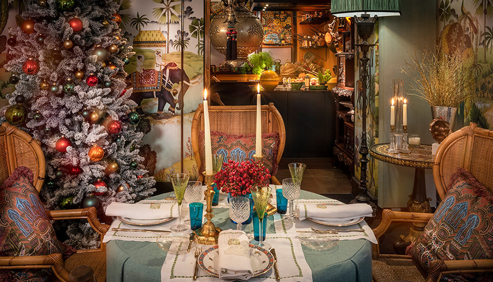 Une table décorée avec des verres colorés et des bougies est placée devant un sapin de Noël enneigé et orné de boules multicolores.