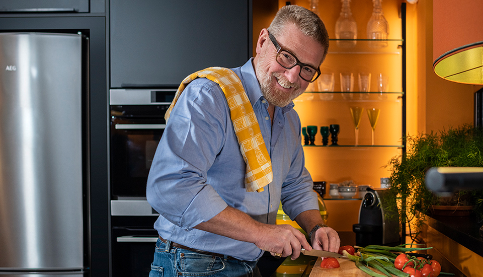Un homme en chemise bleue découpe des légumes sur une planche en bois dans une cuisine moderne avec des étagères en verre.