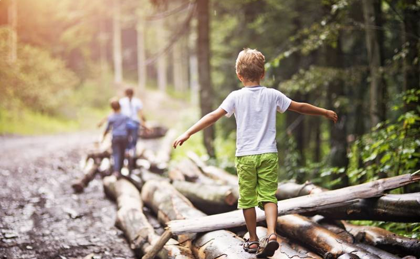 Un enfant en short vert et t-shirt blanc marche en équilibre sur des troncs d'arbres dans une forêt ensoleillée.