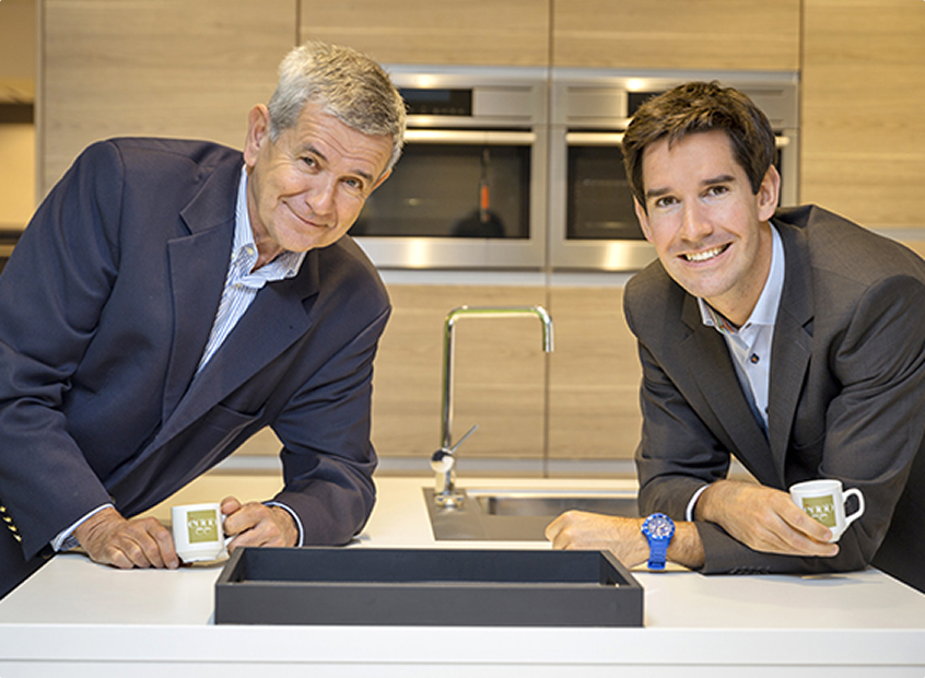 Deux hommes en costume tenant des tasses à café dans une cuisine moderne avec un évier et des appareils en arrière-plan.