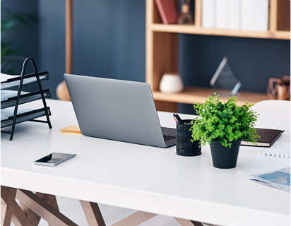 Un ordinateur portable gris est posé sur un bureau blanc, accompagné d'un pot de plante verte, d'un smartphone et de dossiers noirs.