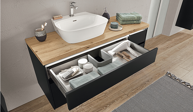 Un meuble-lavabo en bois clair avec vasque blanche, tiroir ouvert contenant serviettes vertes et accessoires de salle de bain, sur sol en bois.