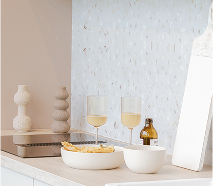 Deux verres à pied remplis de liquide jaune, posés sur un plan de travail avec des bols blancs, une bouteille brune et des objets décoratifs.