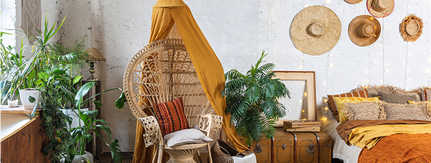 Fauteuil en rotin avec coussins, couvert d'un tissu jaune, entouré de plantes vertes, à côté d'un lit avec des coussins beiges et oranges.