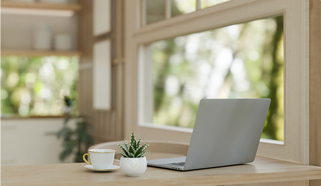 Un ordinateur portable gris, une tasse blanche avec soucoupe jaune et une petite plante en pot sont posés sur une table en bois devant une grande fenêtre.