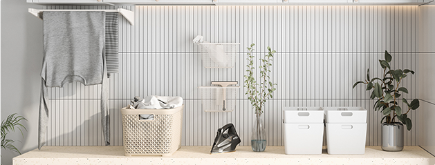 Des serviettes grises sur un porte-serviettes, un panier à linge, un fer à repasser et des plantes sur un plan de travail beige.