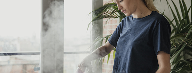 Une personne portant un t-shirt bleu utilise un fer à repasser avec de la vapeur, près d'une fenêtre et de plantes vertes.