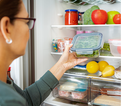 Une personne range un récipient en plastique contenant des légumes verts dans un réfrigérateur rempli de fruits, légumes et condiments.