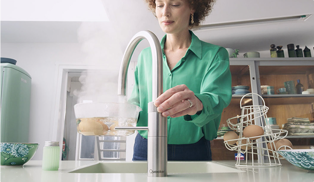 Une femme en chemise verte utilise un robinet moderne dans une cuisine, avec des œufs posés dans un support à côté.