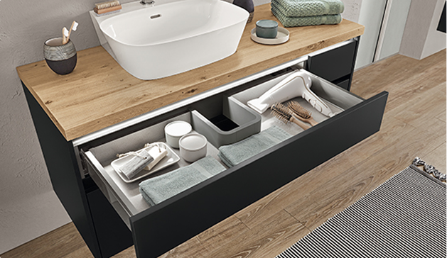Tiroir de salle de bain noir ouvert sous un plan en bois avec lavabo blanc, contenant serviettes, brosses et accessoires de toilette organisés.