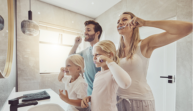 Une famille de quatre personnes se brosse les dents devant un miroir dans une salle de bain lumineuse.