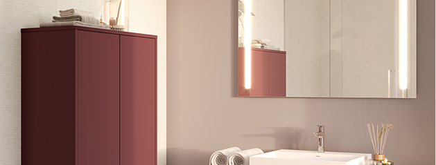 Un meuble de salle de bain bordeaux à gauche et un lavabo blanc avec un miroir éclairé à droite.