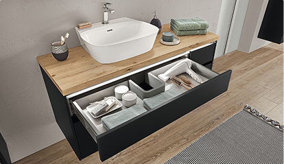 Un meuble de salle de bain en bois clair avec lavabo blanc, tiroir ouvert contenant serviettes vertes et accessoires, situé sur un sol en bois.