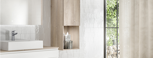 Un lavabo blanc avec un robinet chromé est installé sur un meuble en bois clair, à côté de carreaux blancs et d'une fenêtre.