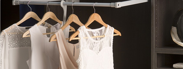 Des vêtements blancs et beiges sont suspendus sur des cintres en bois dans une armoire sombre, à côté de compartiments de rangement.