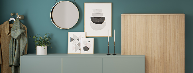 Un meuble vert avec des cadres, un miroir rond, deux bougies noires et une plante en pot sur fond de mur bleu.