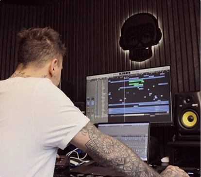 Un homme tatoué en t-shirt blanc travaille sur un projet audio sur deux écrans d'ordinateur dans un studio sombre.