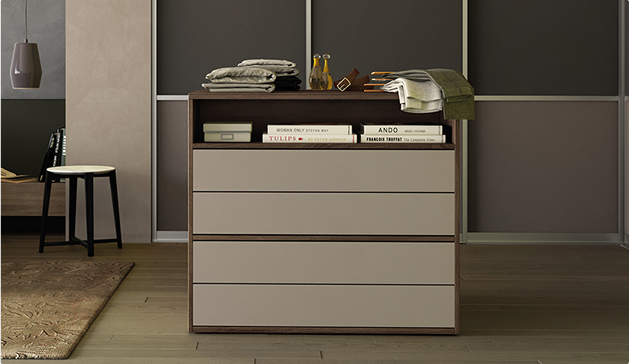 Commode moderne beige et marron avec des livres, bouteilles et textiles sur le dessus, dans une pièce au sol en bois et murs sombres.