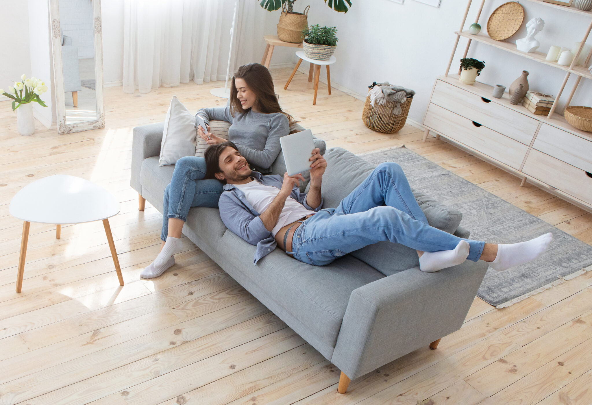 Un couple détendu est assis sur un canapé gris dans un salon lumineux avec un sol en bois clair et une table basse blanche.